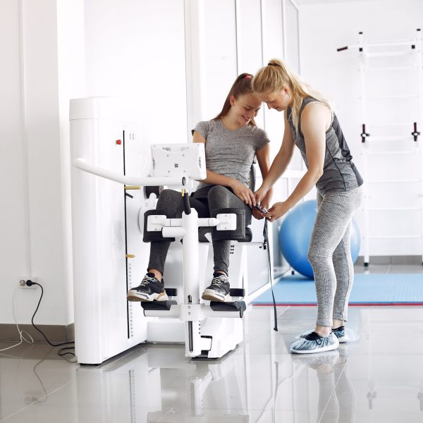 A therapist is coaching the girl. The patient is doing the exercises. The women are in the gym.